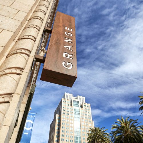 Sunlit city street view of tall columns and a rust-colored sign with bold letters against a blue sky.