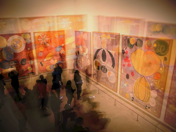 A museum gallery with bright abstract artworks and visitors standing along the wall, admiring colorful paintings.