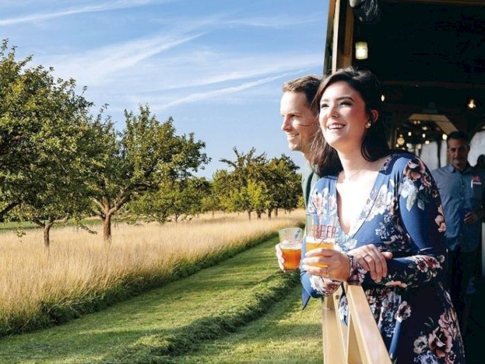 Two women smile with beer in hand on a sunny garden patio by an open vehicle, with trees and a grassy orchard behind.