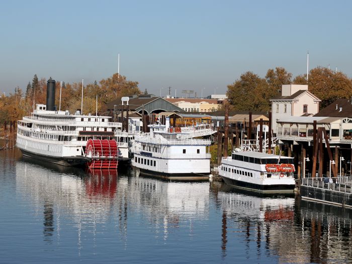 Two white river cruise ships are docked by wooden piers along a calm river, with trees and a U.S. flag flying.
