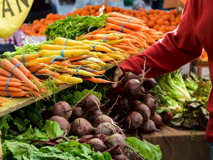 A farmer&rsquo;s market stall piled with carrots and leafy greens, with an arm reaching in to pick veggies.