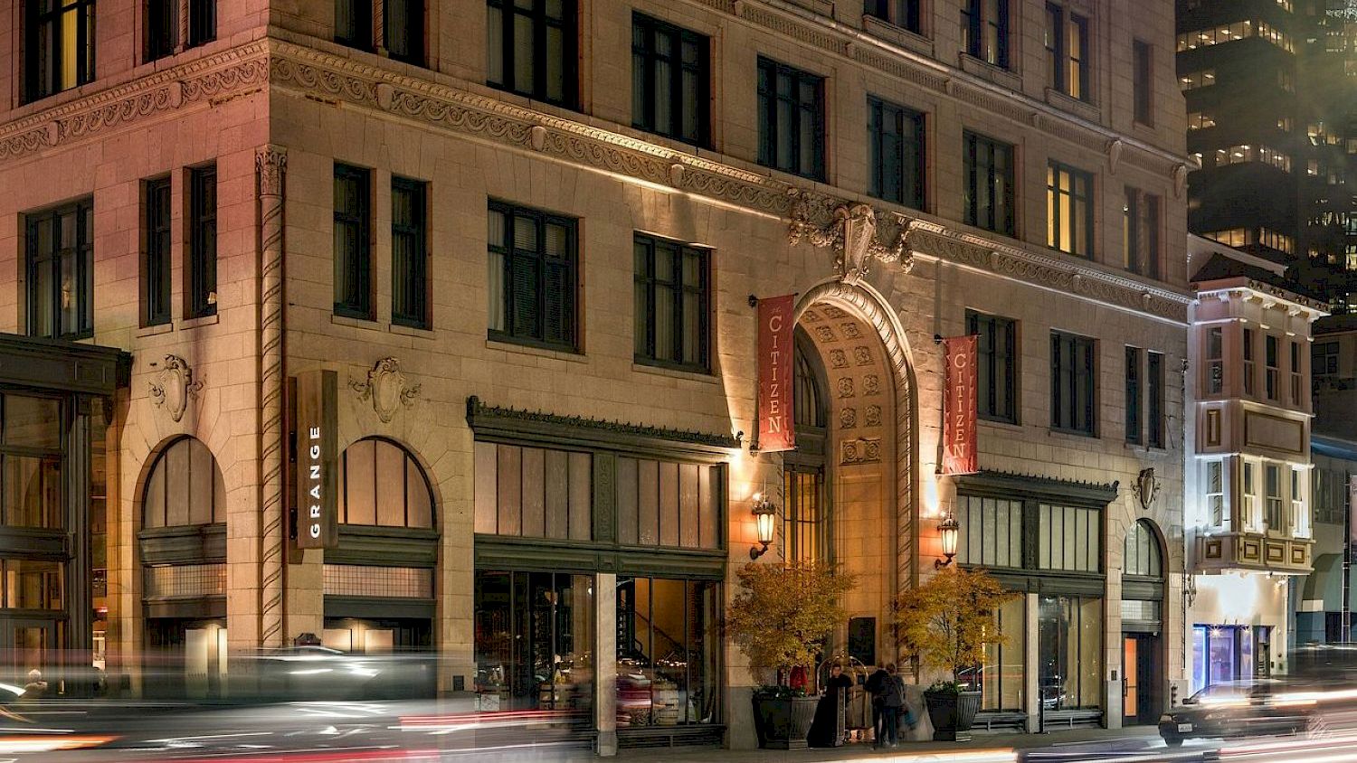 Historic stone building at night with vertical &ldquo;Cityzen&rdquo; banners and light-trail traffic streaks rushing past, in a city street scene.