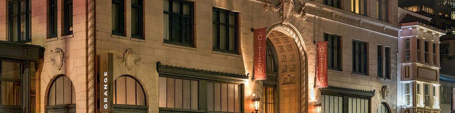 Historic stone building at night with vertical &ldquo;Cityzen&rdquo; banners and light-trail traffic streaks rushing past, in a city street scene.