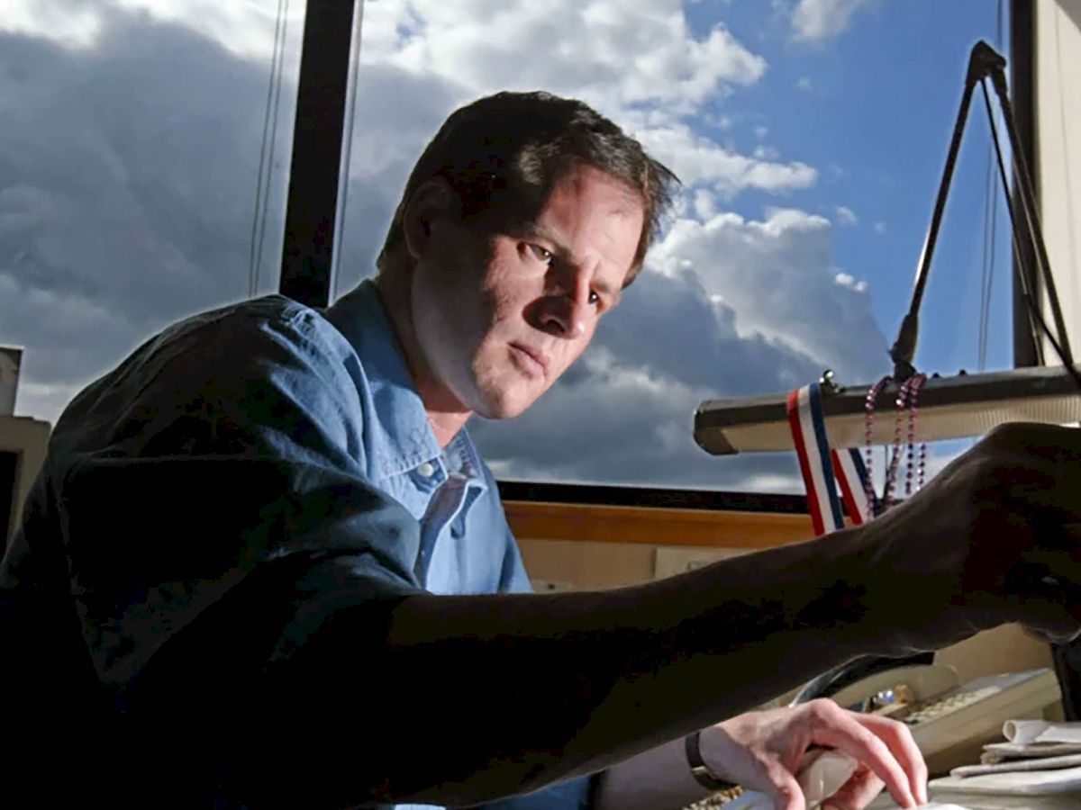 A man in a workshop leans over a surface with tools while looking toward a window with clouds outside.