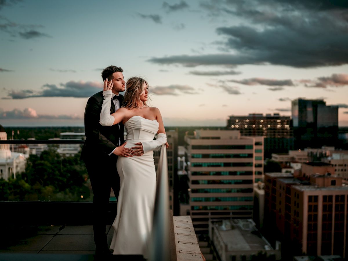 A bride and groom embrace on a rooftop at sunset, gazing over a city skyline and swirling clouds.