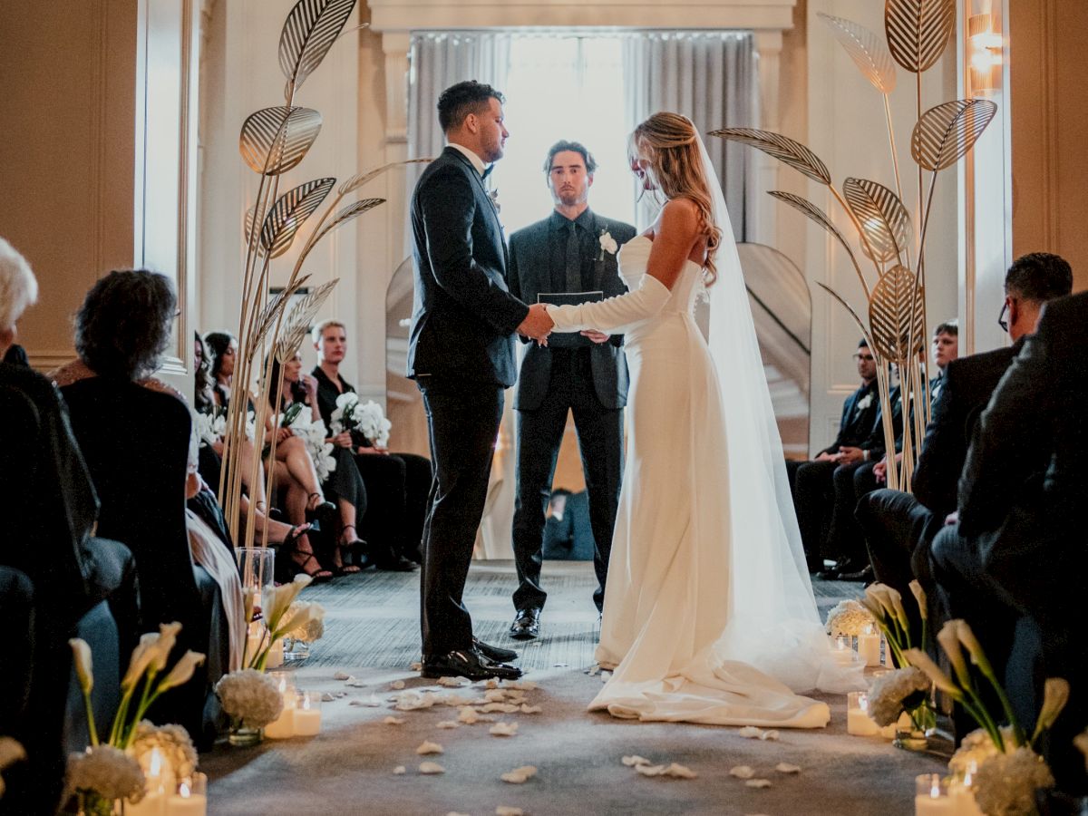 A couple exchanges vows at an elegant indoor wedding ceremony, with guests seated and a chandelier overhead.