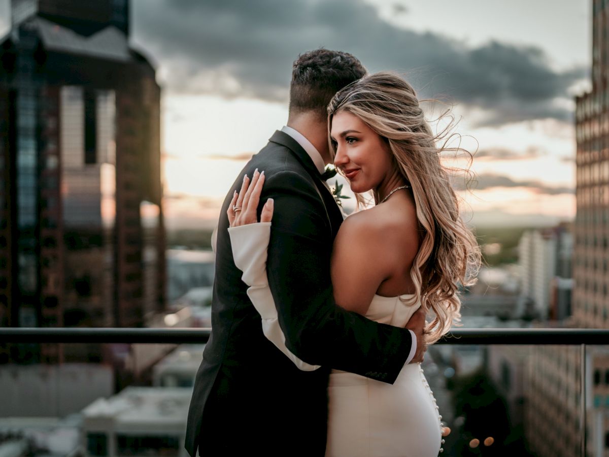 A bride and groom in formalwear share a romantic embrace on a city rooftop at sunset, smiling as vows begin.