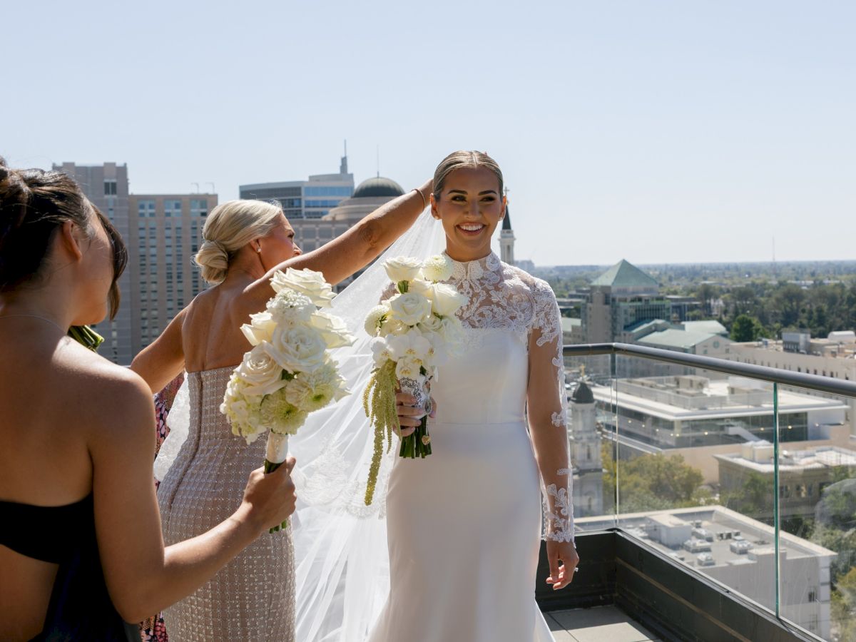 A bride in a white gown smiles on a balcony as friends adjust her veil, holding white bouquets overlooking the city.
