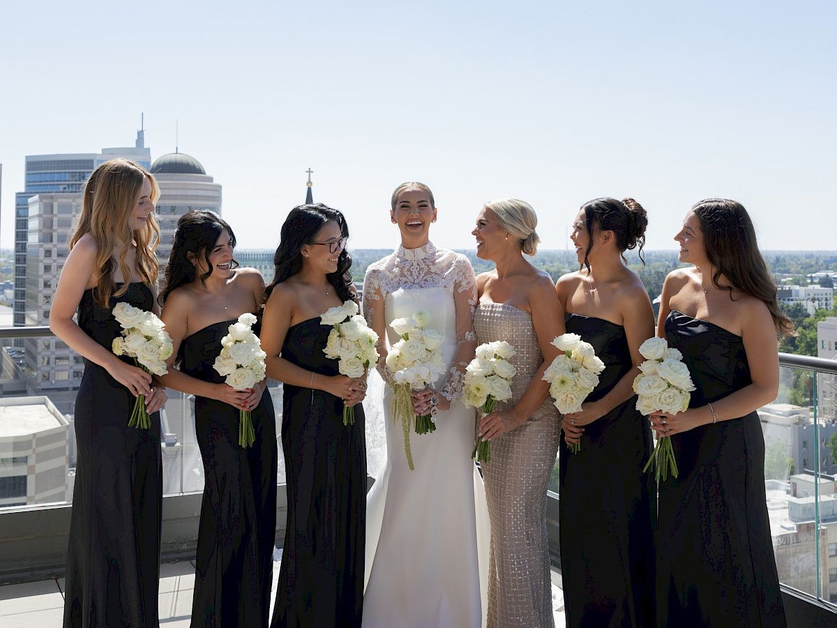 A bride and her bridesmaids in evening dresses smile on a rooftop, holding white rose bouquets against a city skyline.