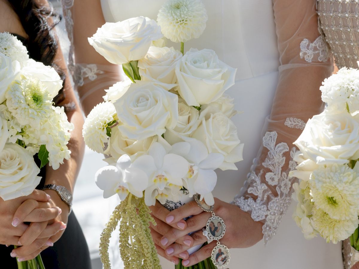 A bride and groom in elegant attire hold white wedding bouquets with delicate lace sleeves and gold-toned flower accents.