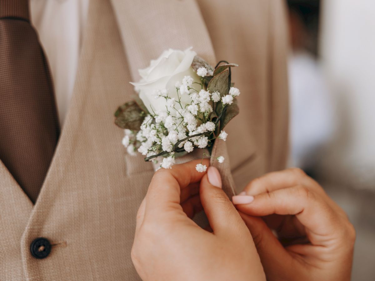 A hand pins a small white flower boutonniere onto a tan suit lapel, with soft light and a gentle wedding vibe.