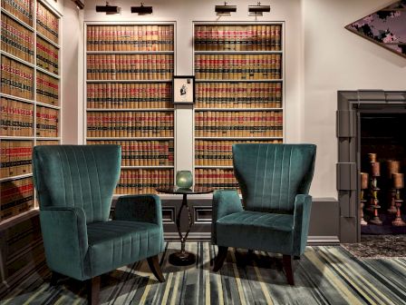 A cozy office/library with tall shelves of old books, teal armchairs, a patterned carpet, and a small framed artwork.